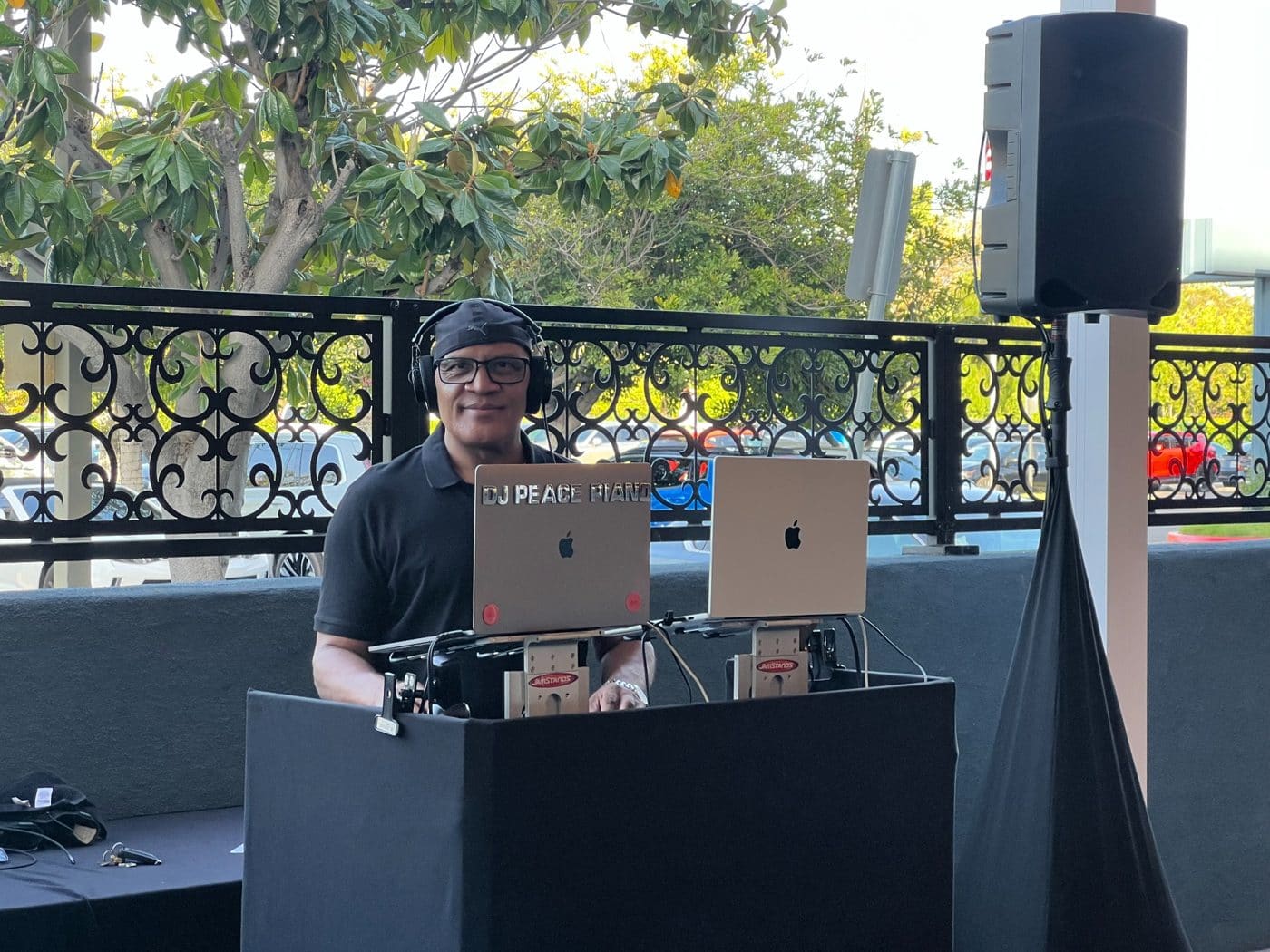 DJ at outdoor event with dual laptops and mixing equipment behind ornate black railing