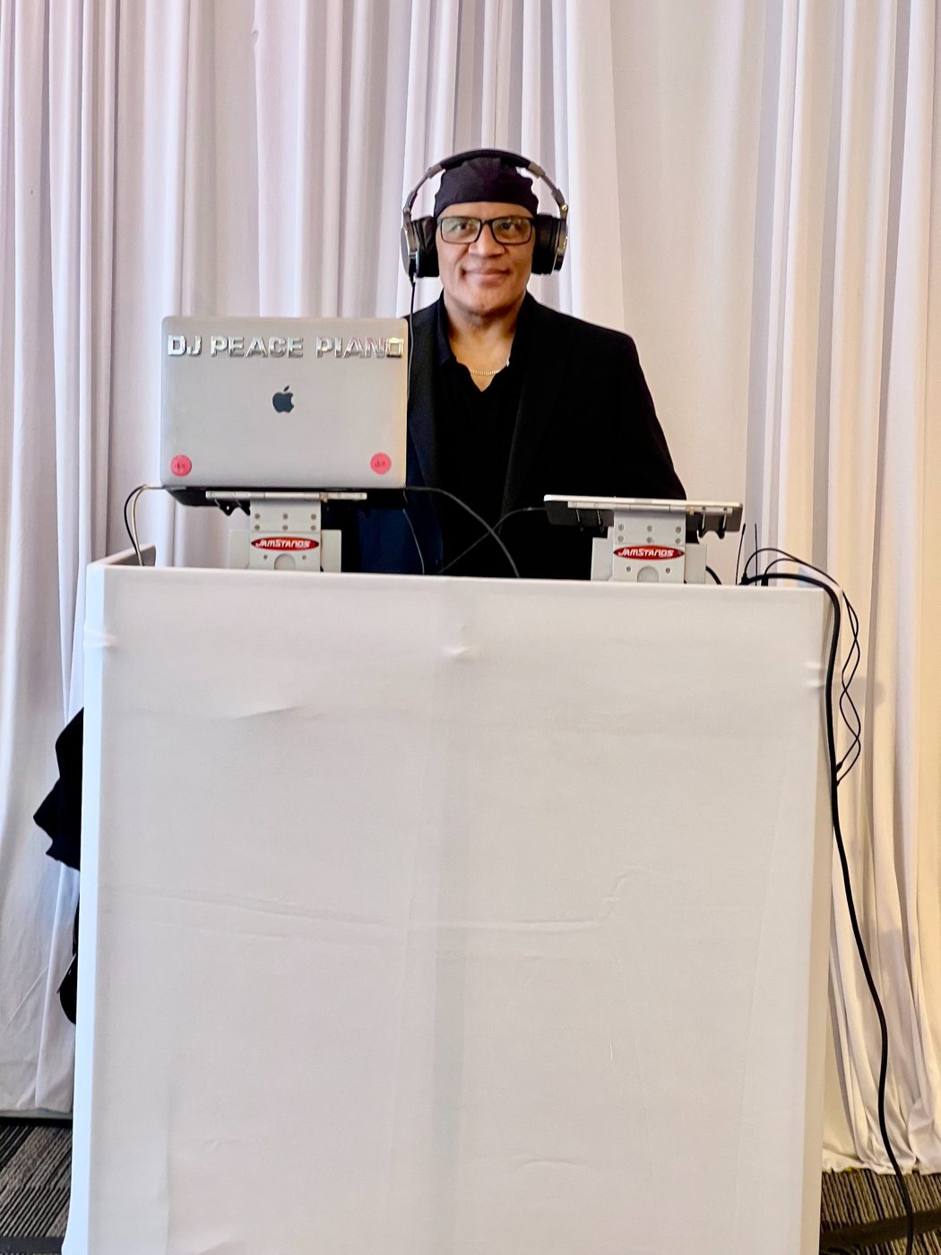 DJ Peace Piang at turntables wearing headphones and glasses behind white booth