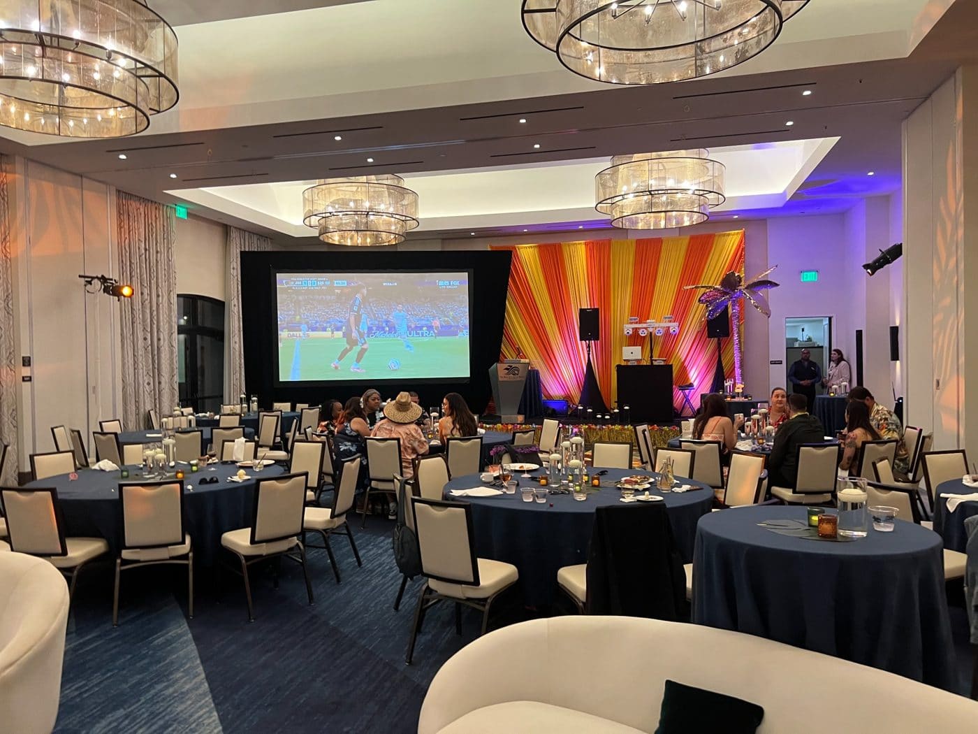 Elegant ballroom event space with round tables, projection screen showing soccer match, and colorful stage lighting