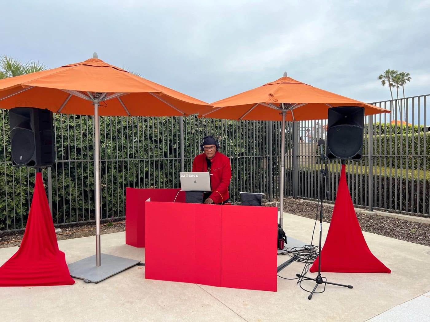 DJ at outdoor event setup with red booth, orange umbrellas and palm trees