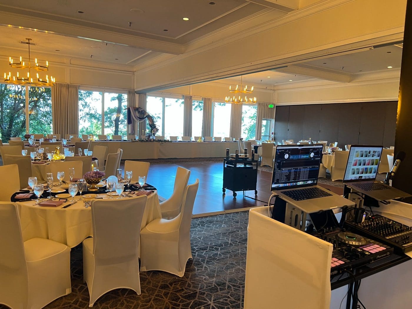 Elegant ballroom setup with round dining tables, white chair covers, floor-to-ceiling windows, stage with blue lighting, and DJ equipment